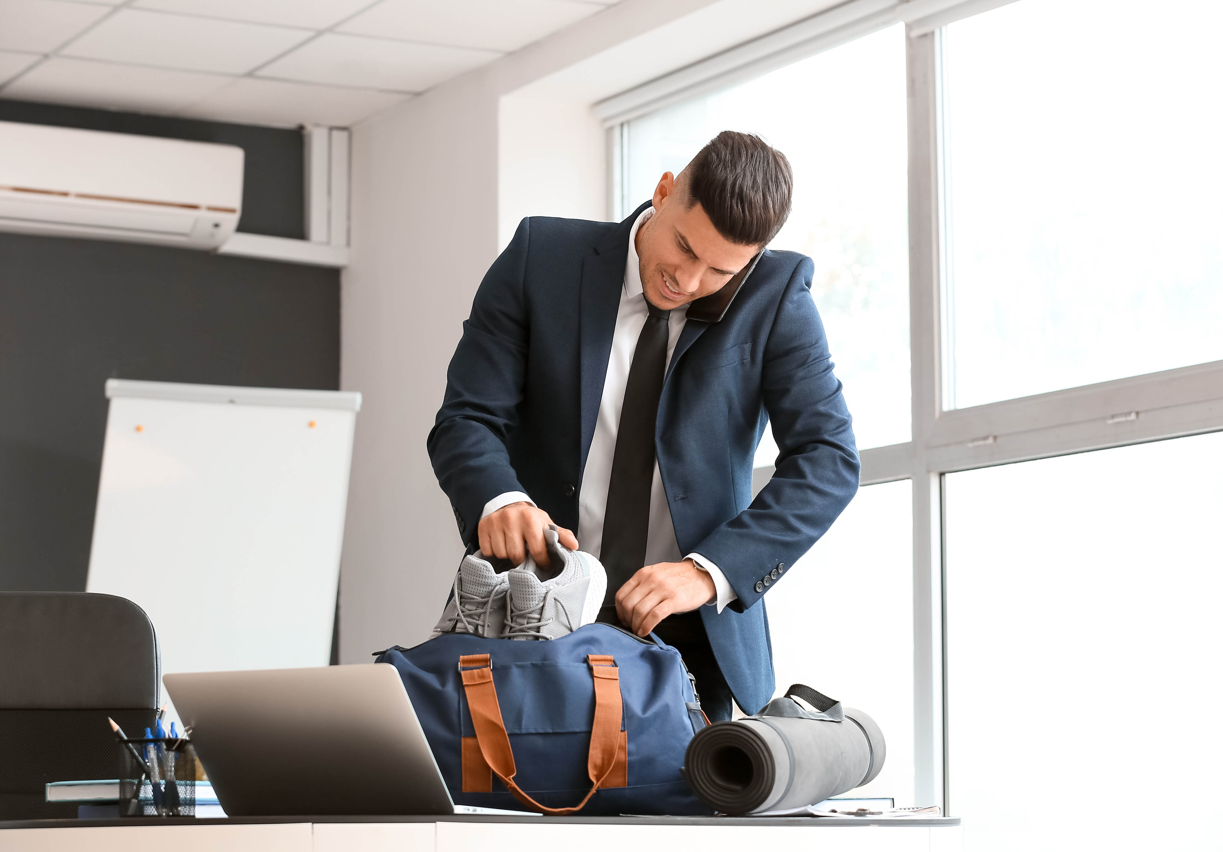 Young business professional on the phone in the office packing his shoes for a workout at the gym